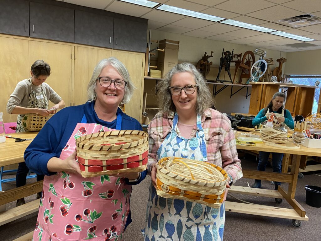 Two women stand side by side holding completed reed baskets