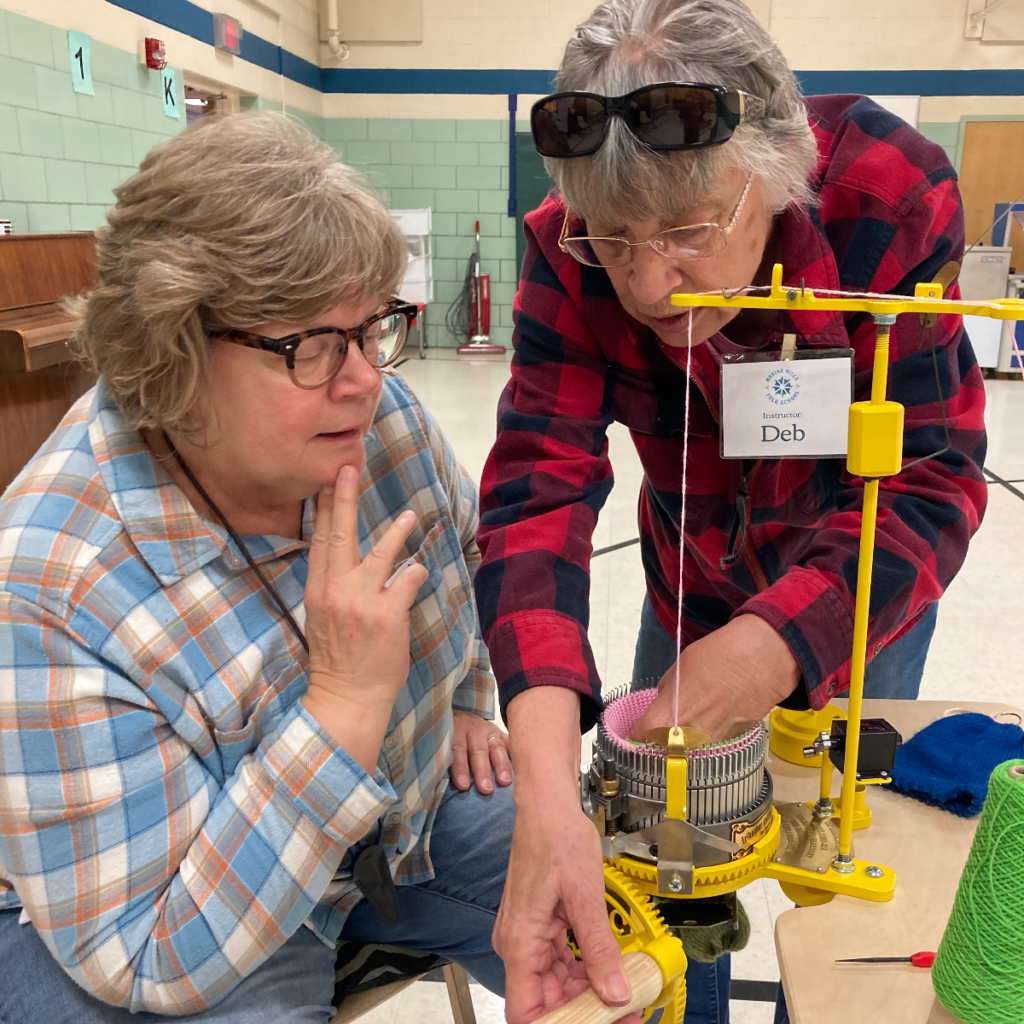 A woman demonstrates how to use a sock crank machine to another woman.